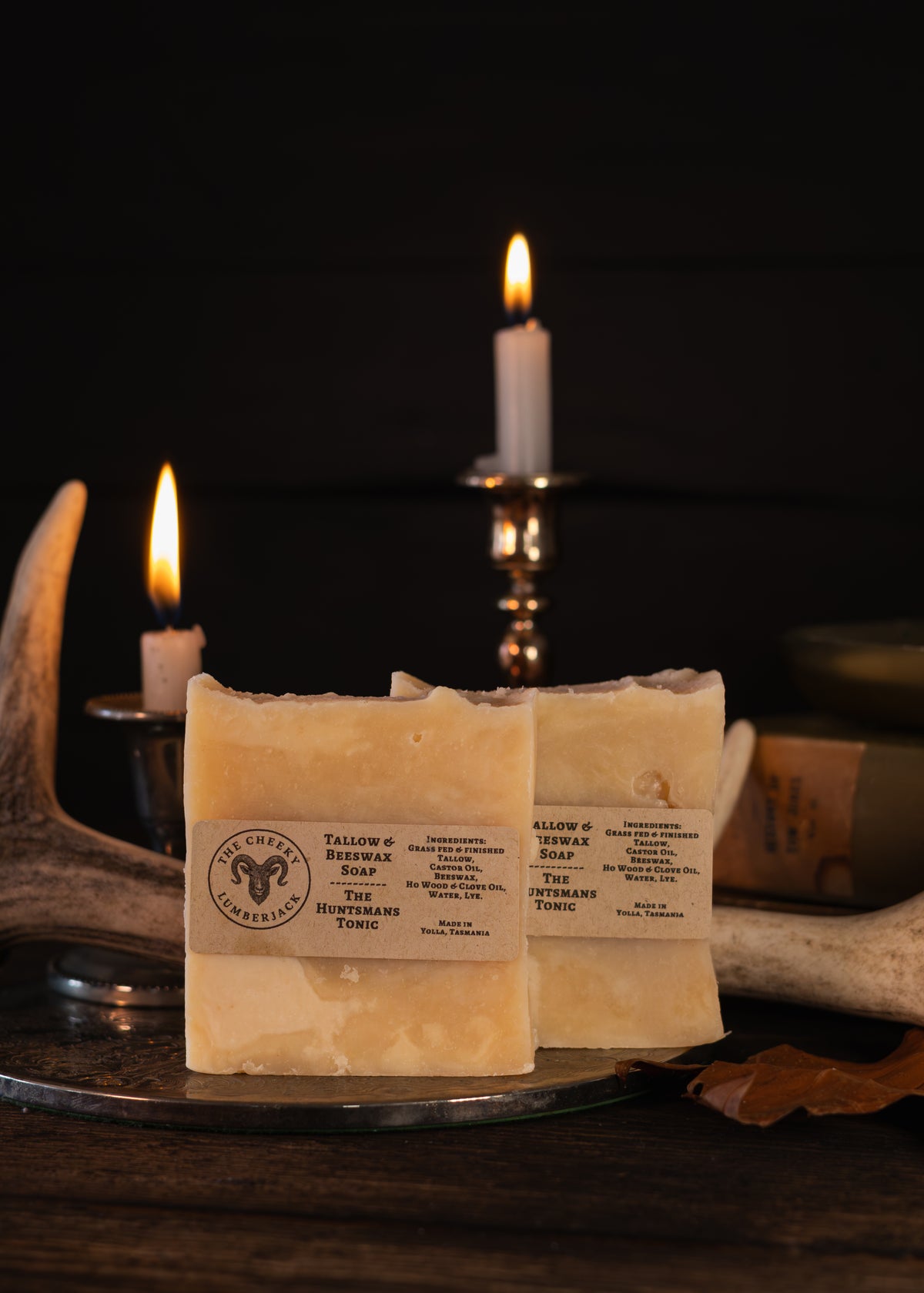 Two bars of handmade soap labeled 'The Huntsman's Tonic' resting on a metal tray, styled with lit candles, antlers, and a dark rustic background.