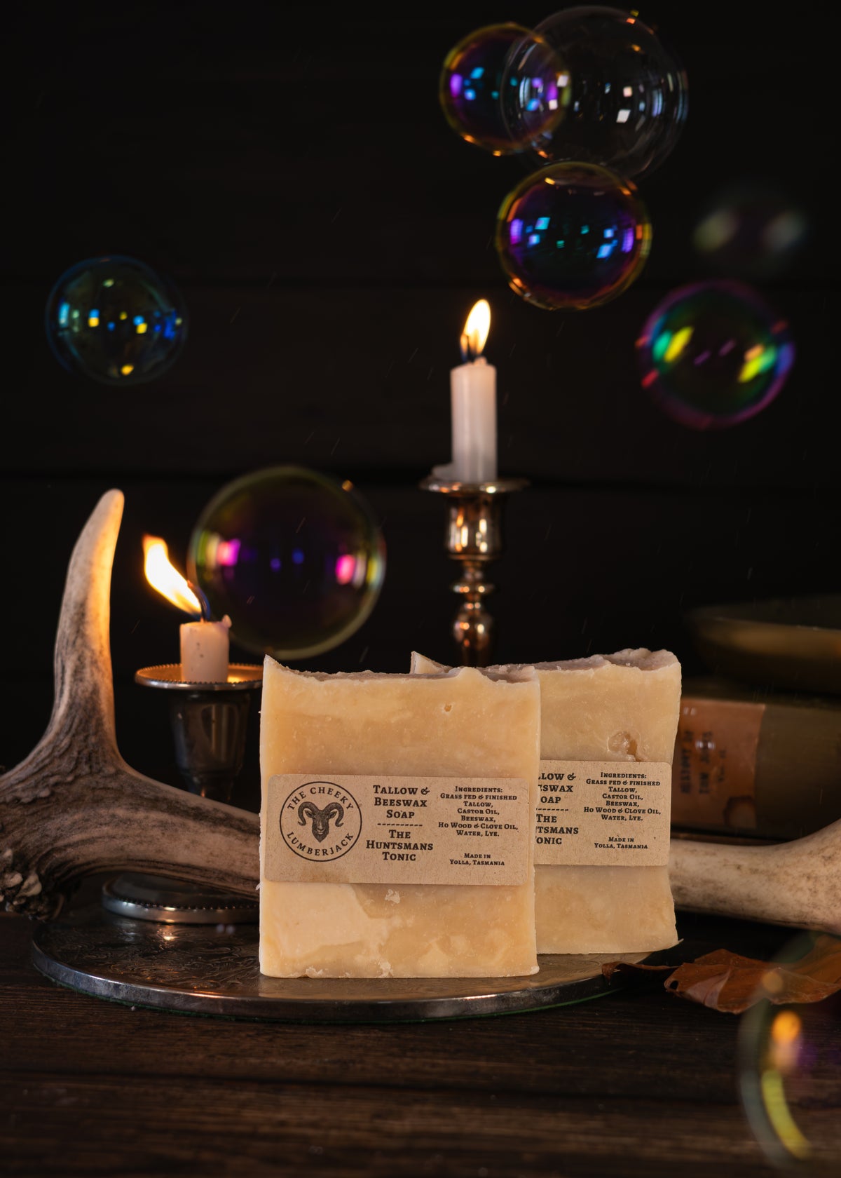 Two bars of Salting the Earth Soap, 'The Cheeky Lumberjack' displayed on a rustic metal tray with candles, antlers, and floating bubbles against a dark background.