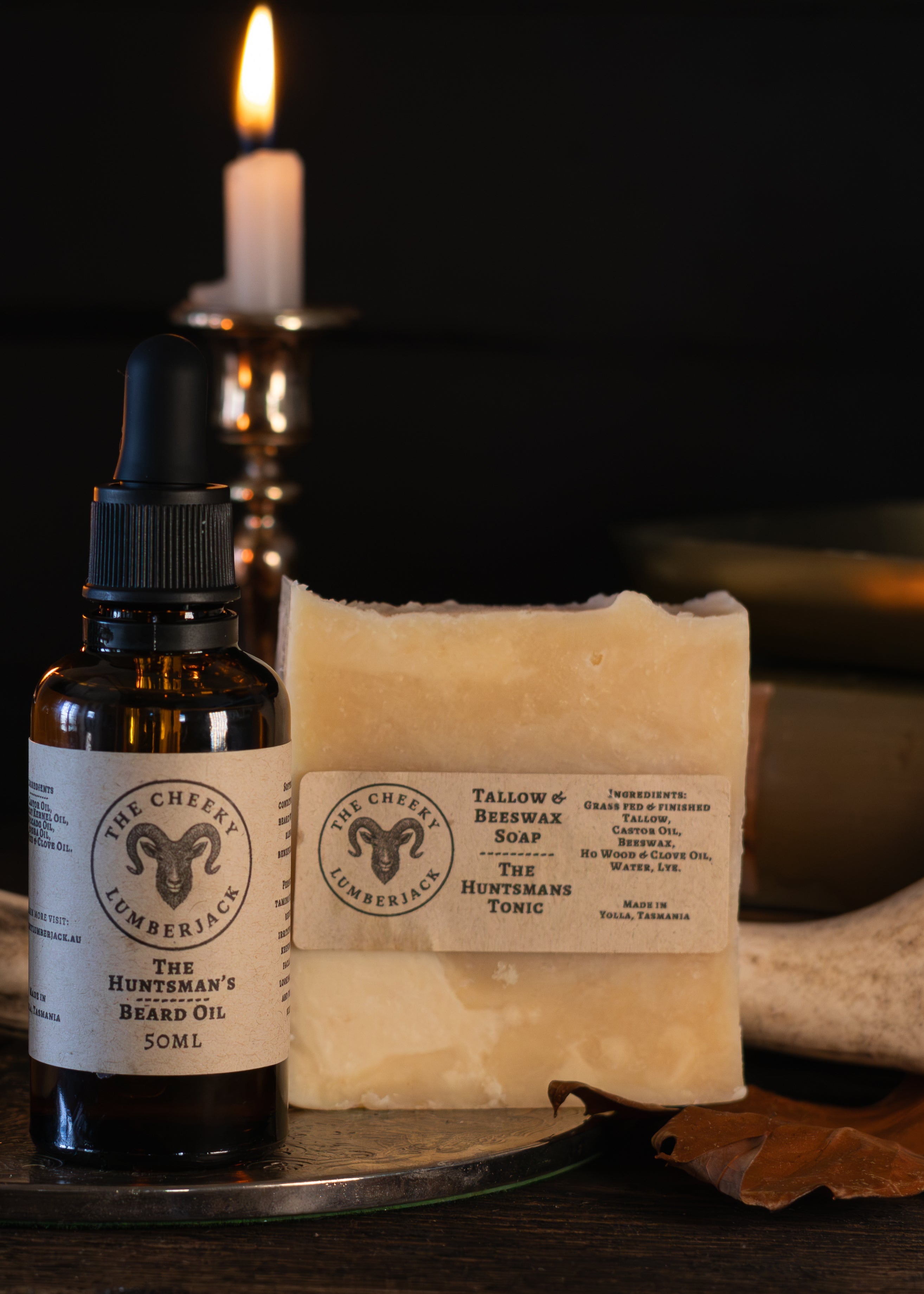 Three artisanal products from Wild Earth Apothecary displayed on a glass tray: a jar of The Apothecary Salve, a bottle of Bittern's Balm 50ml, and a bar of Wallow of Wisdom Handmade soap. The products are styled with lit candles, antlers, and a leaf on a dark background.