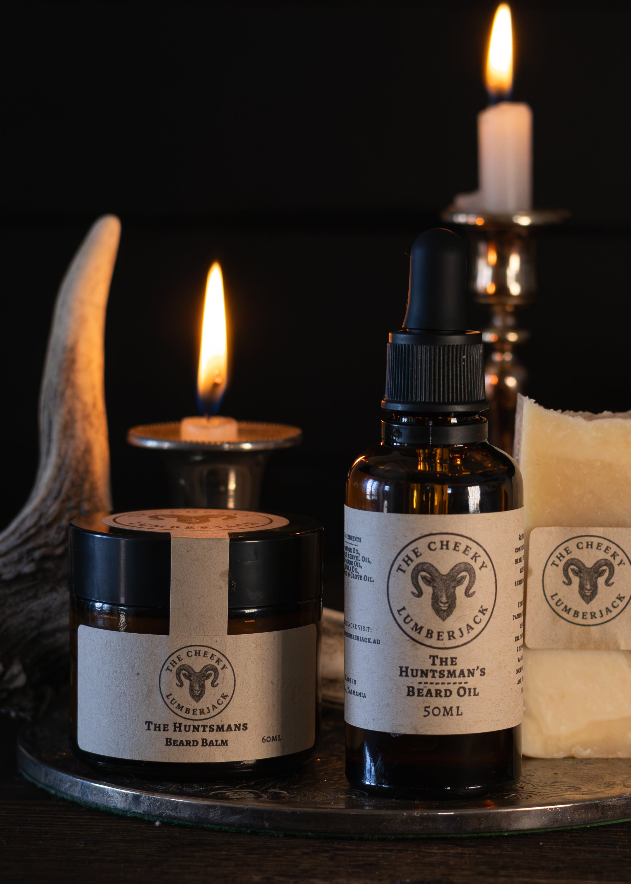 Three artisanal products from Wild Earth Apothecary displayed on a glass tray: a jar of The Apothecary Salve, a bottle of Bittern's Balm 50ml, and a bar of Wallow of Wisdom Handmade soap. The products are styled with lit candles, antlers, and a leaf on a dark background.