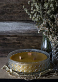 Decorative candle with intricate design on a textured surface with a vase of flowers in the background.