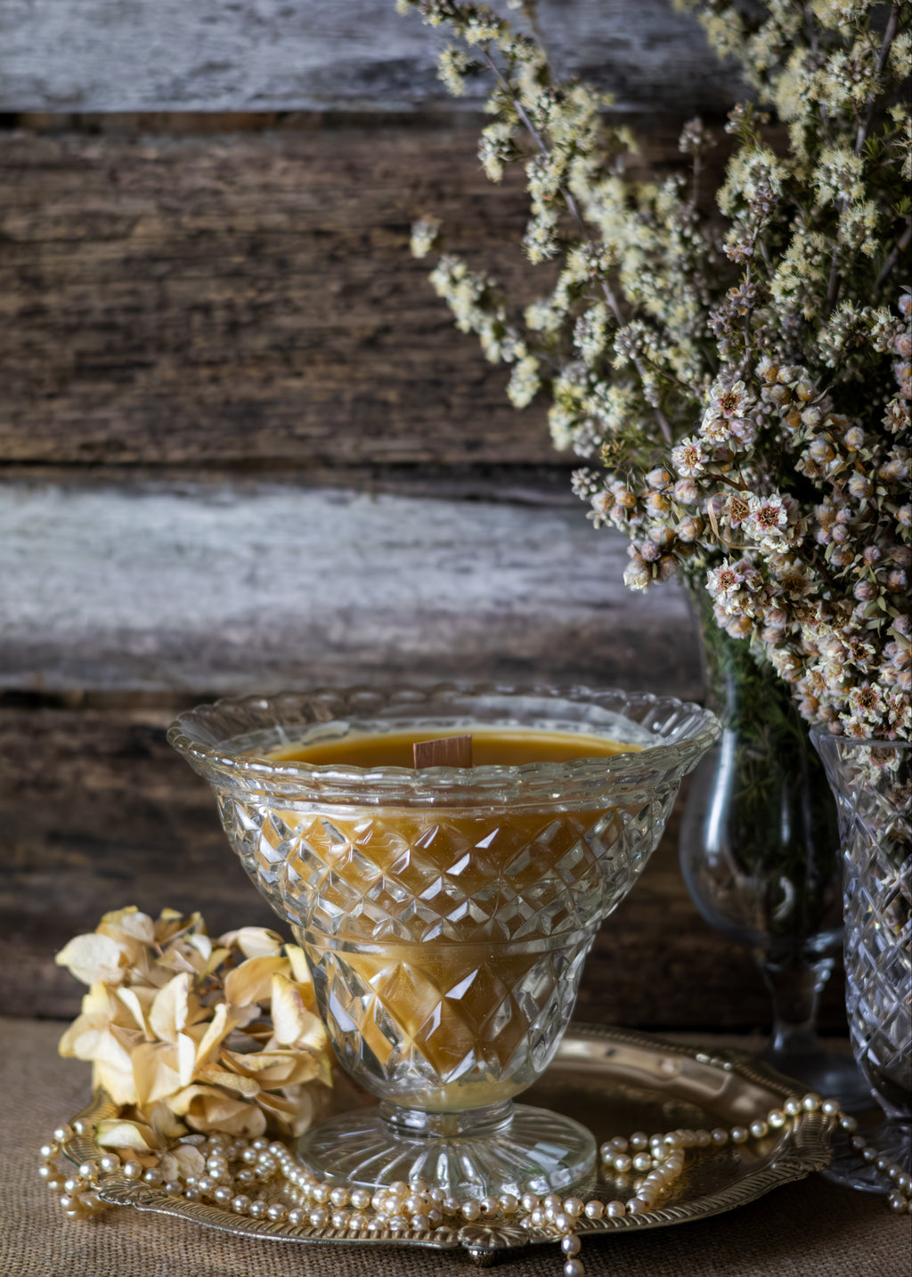 Decorative glass bowl with candle on a rustic wooden surface with flowers and pearls.
