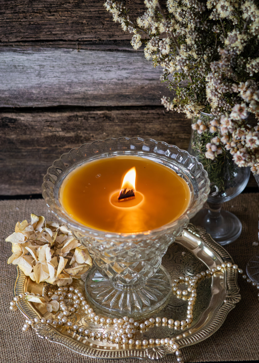 Candle in a glass holder on a decorative tray with flowers and dried elements against a wooden background