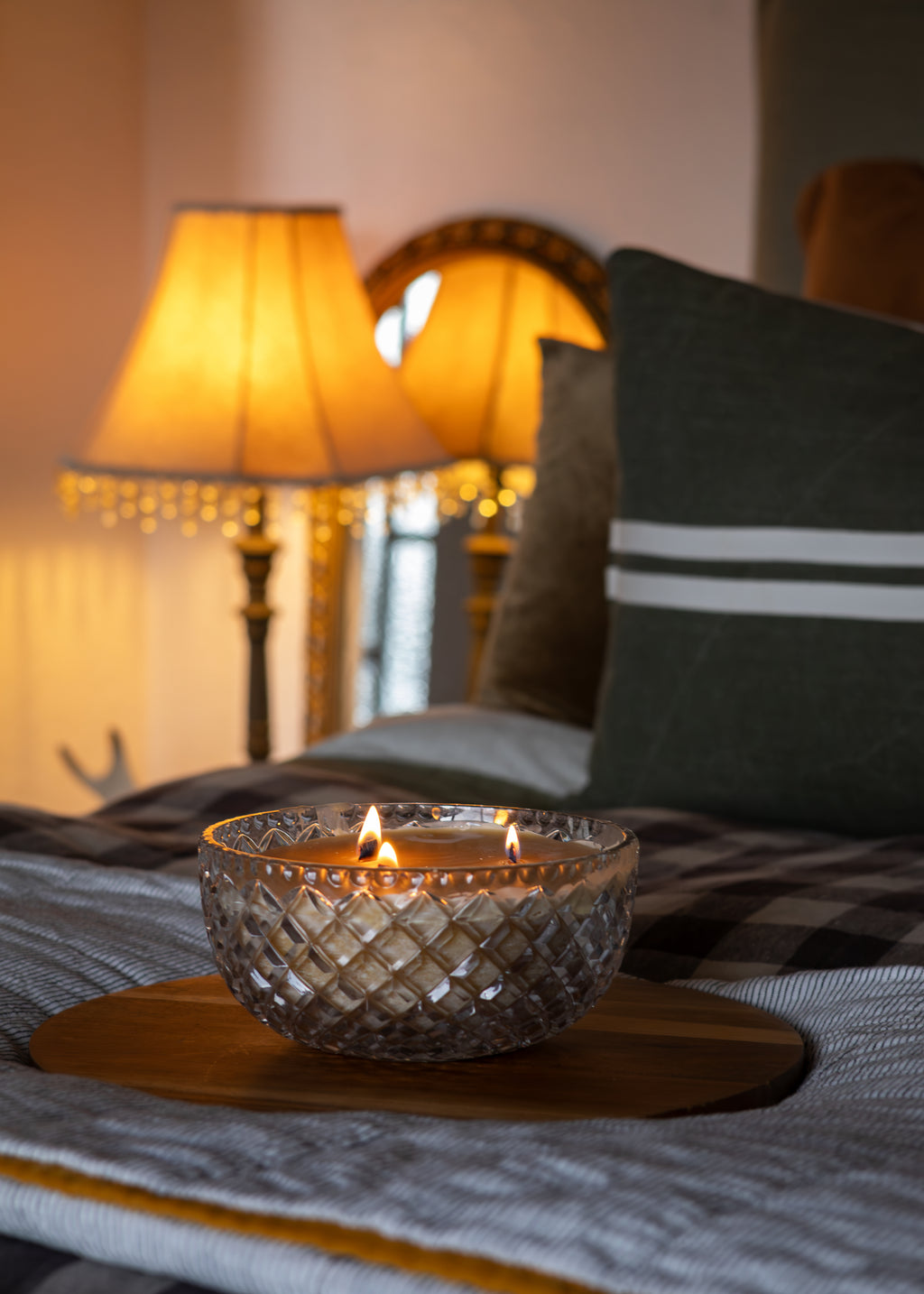 A lit two-wick candle in a clear cut-crystal textured bowl sits on a round wooden tray on a bed with a plaid blanket, flanked by a dark green pillow with white stripes and warm glowing bedside lamps in the background.