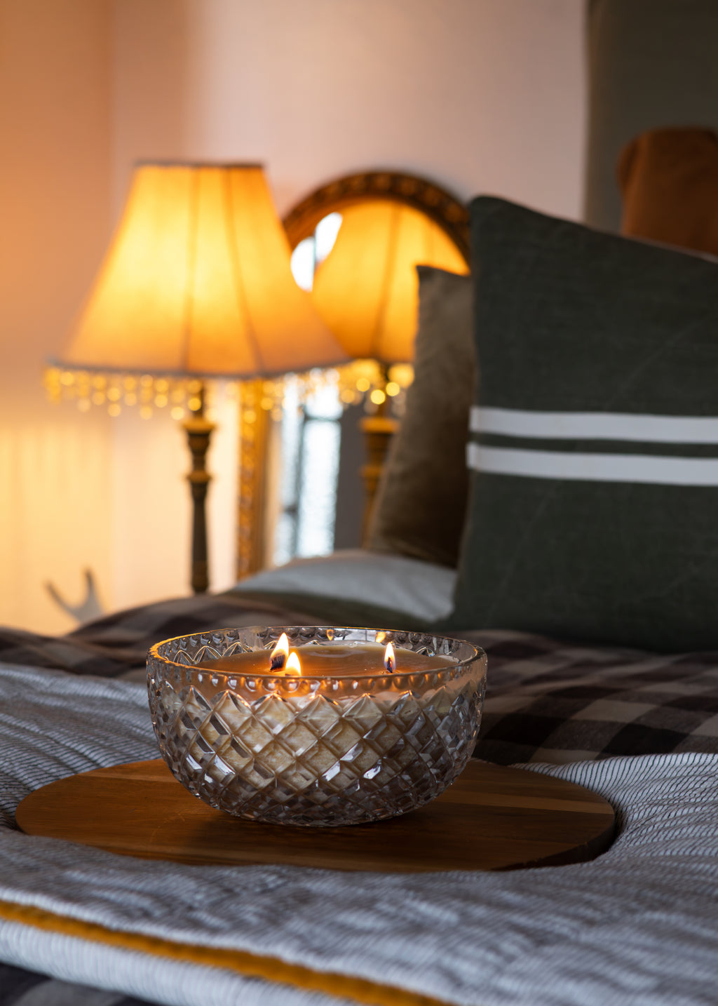 A lit candle in a clear textured glass bowl sits on a round wooden tray on a bed, with a dark green pillow, plaid blanket, and warm glowing bedside lamps in the background.