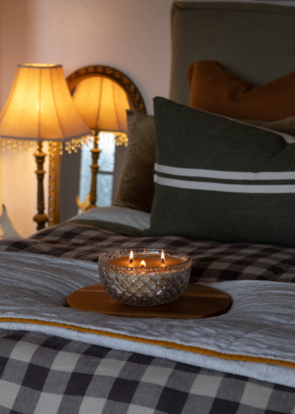 A lit double candle in a textured clear glass bowl sits on a wooden tray on a bed with a gray and white buffalo plaid blanket, green pillows, and warm bedside lamps in the background.
