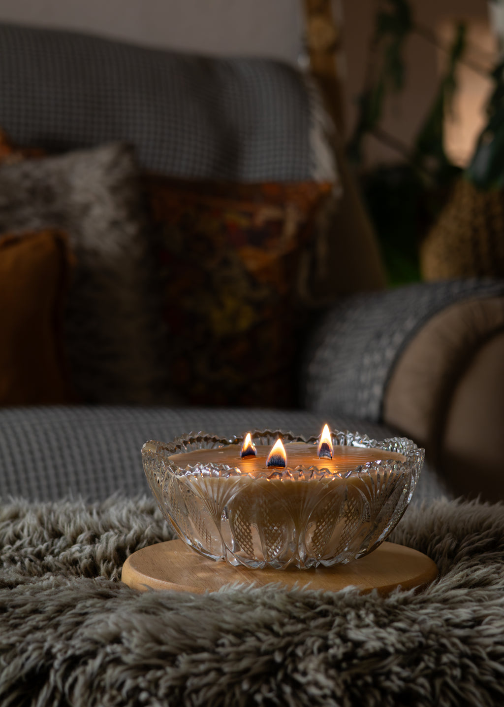 A three-wick glass candle with lit flames sits on a round wooden coaster atop a shaggy gray rug in a cozy living room setting with a sofa and cushions in the background.