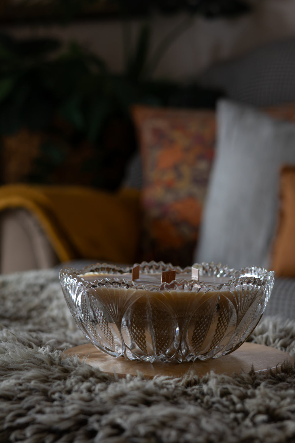 A clear glass bowl with a scalloped rim and textured cut patterns, filled with a light brown candle, resting on a round wooden coaster over a shaggy gray rug in a cozy living room setting.