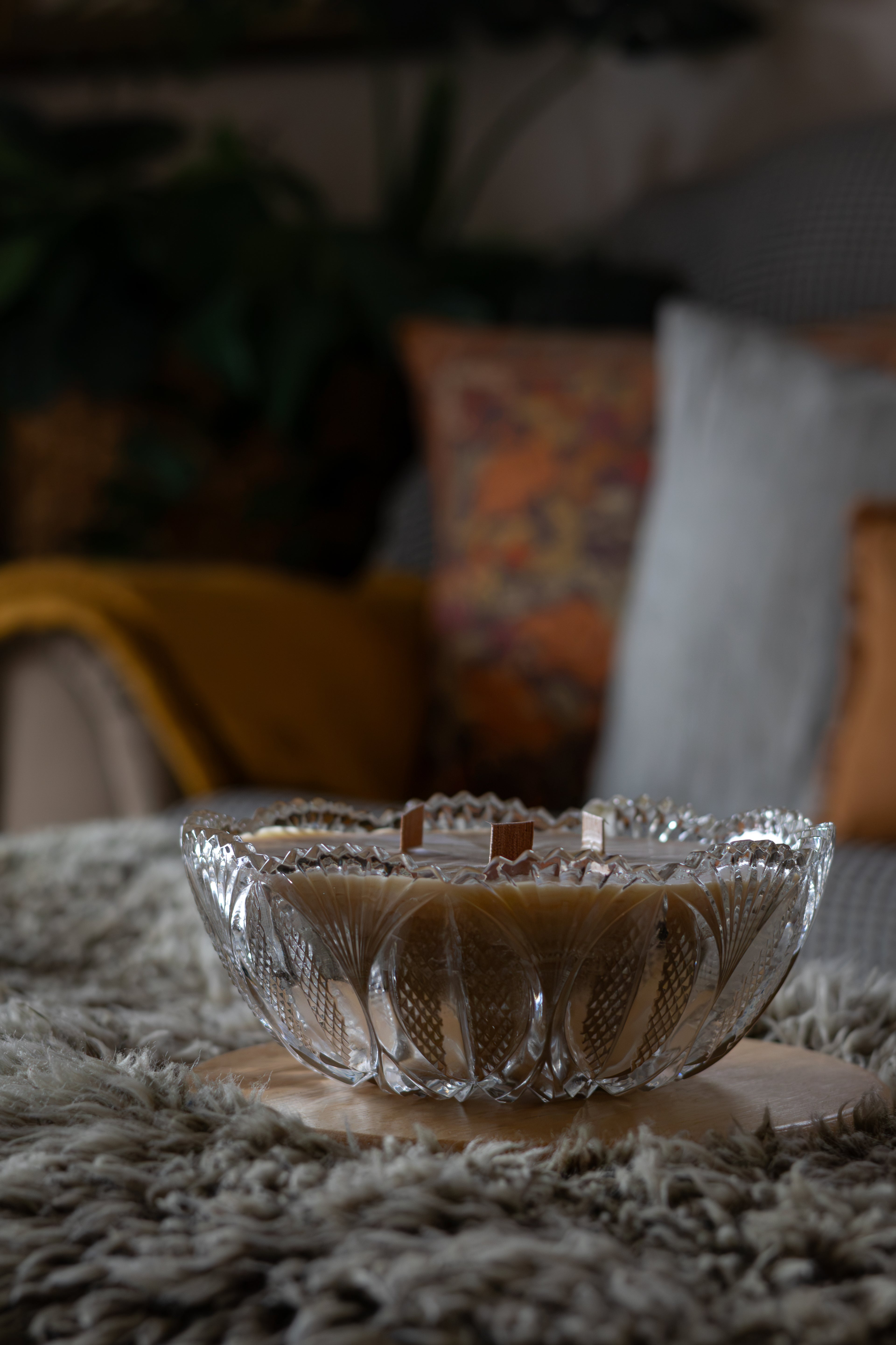 A clear glass bowl with a scalloped rim and textured cut patterns, filled with a light brown candle, resting on a round wooden coaster over a shaggy gray rug in a cozy living room setting.