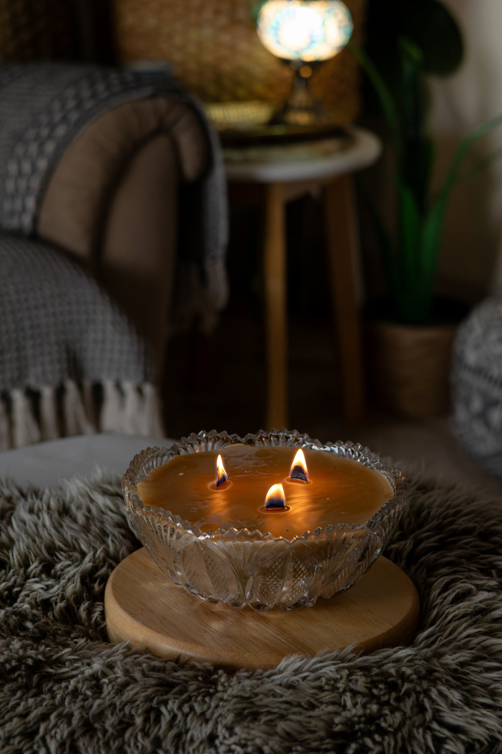 A three-wick candle in a clear textured glass bowl sits on a round wooden tray, lit with three flames in a cozy, dimly lit living room setting.