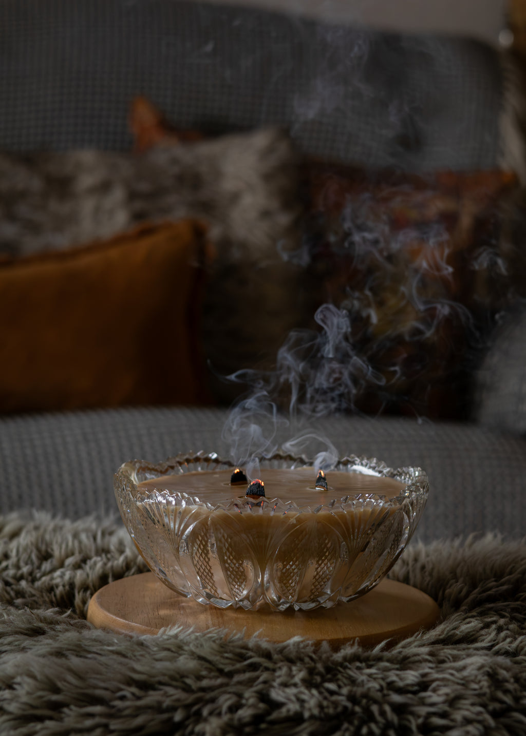 A clear textured glass bowl filled with a light brown liquid, set on a round wooden coaster over a shaggy gray rug. Small burning incense sticks are placed in the center, with visible smoke rising against a blurred living room background with cushions.