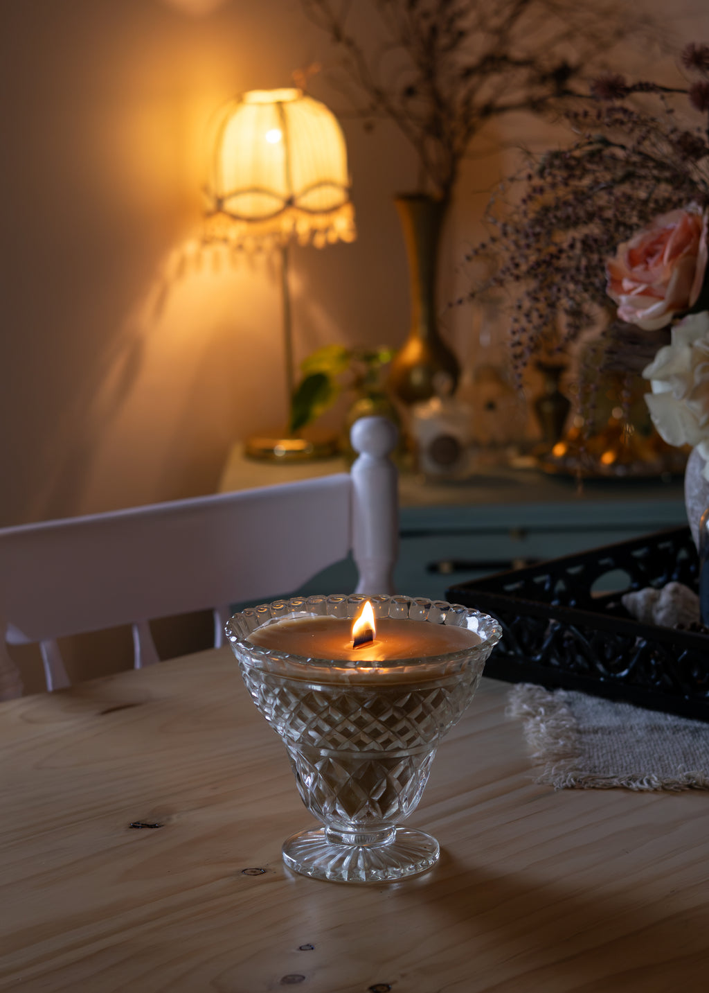 A lit wax candle in a clear cut-glass pedestal holder sits on a wooden table in a warmly lit room, with a lamp, chair, and decorative flowers blurred in the background.