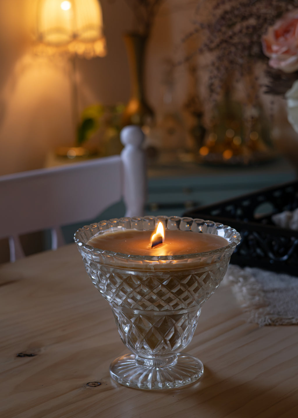 A lit candle in a clear cut-glass pedestal holder sits on a wooden table in a warmly lit, softly blurred room with a lamp and decorative items in the background.
