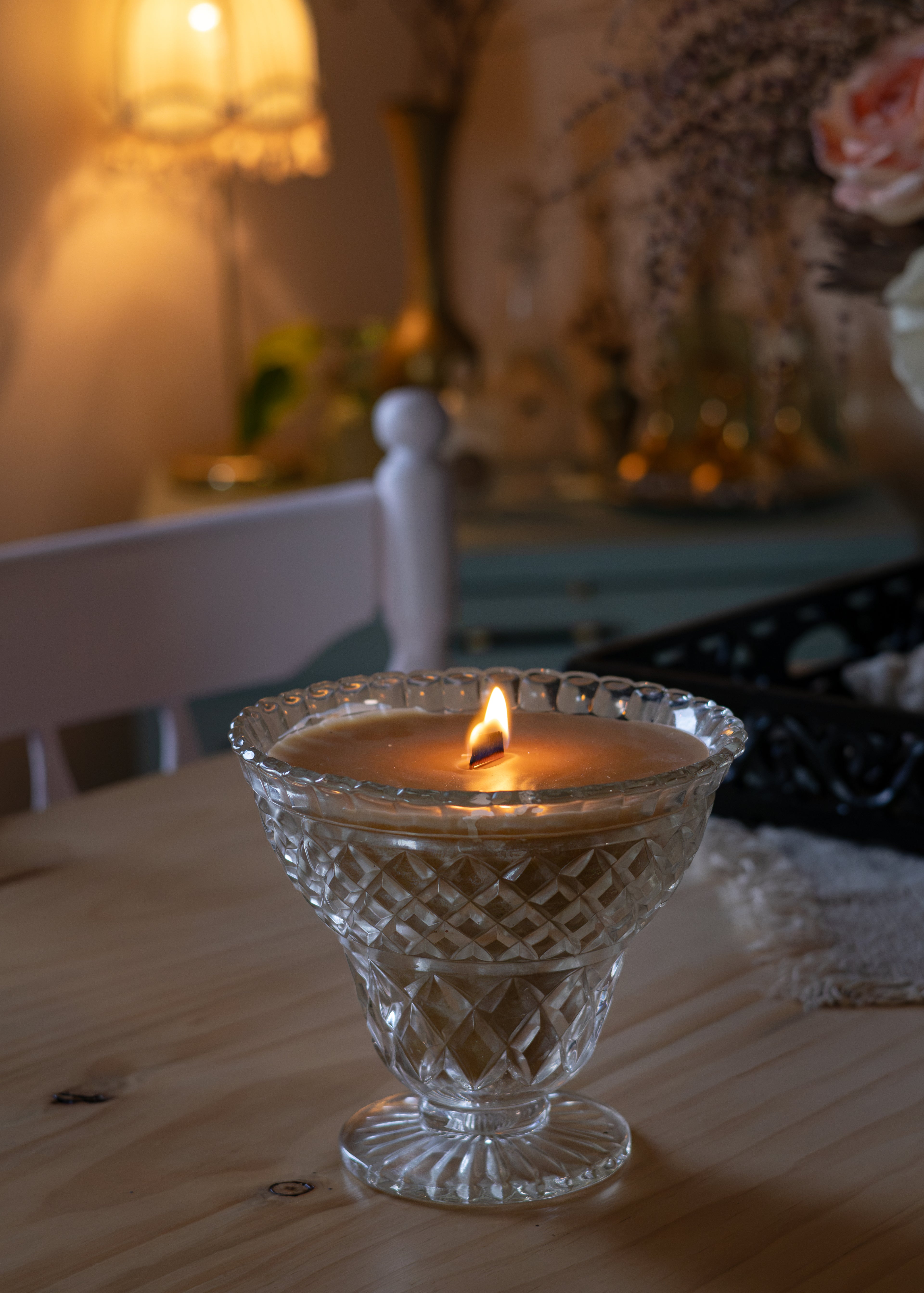 A lit candle in a clear cut-glass pedestal holder sits on a wooden table in a warmly lit, softly blurred room with a lamp and decorative items in the background.