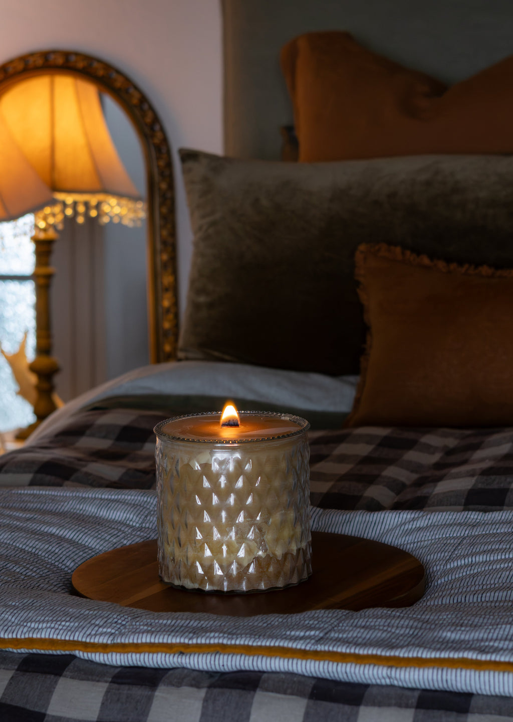 A lit textured glass candle holder with a geometric pattern sits on a wooden coaster on a bed with a gray and white buffalo plaid blanket, styled with brown and gray pillows in a cozy bedroom setting.