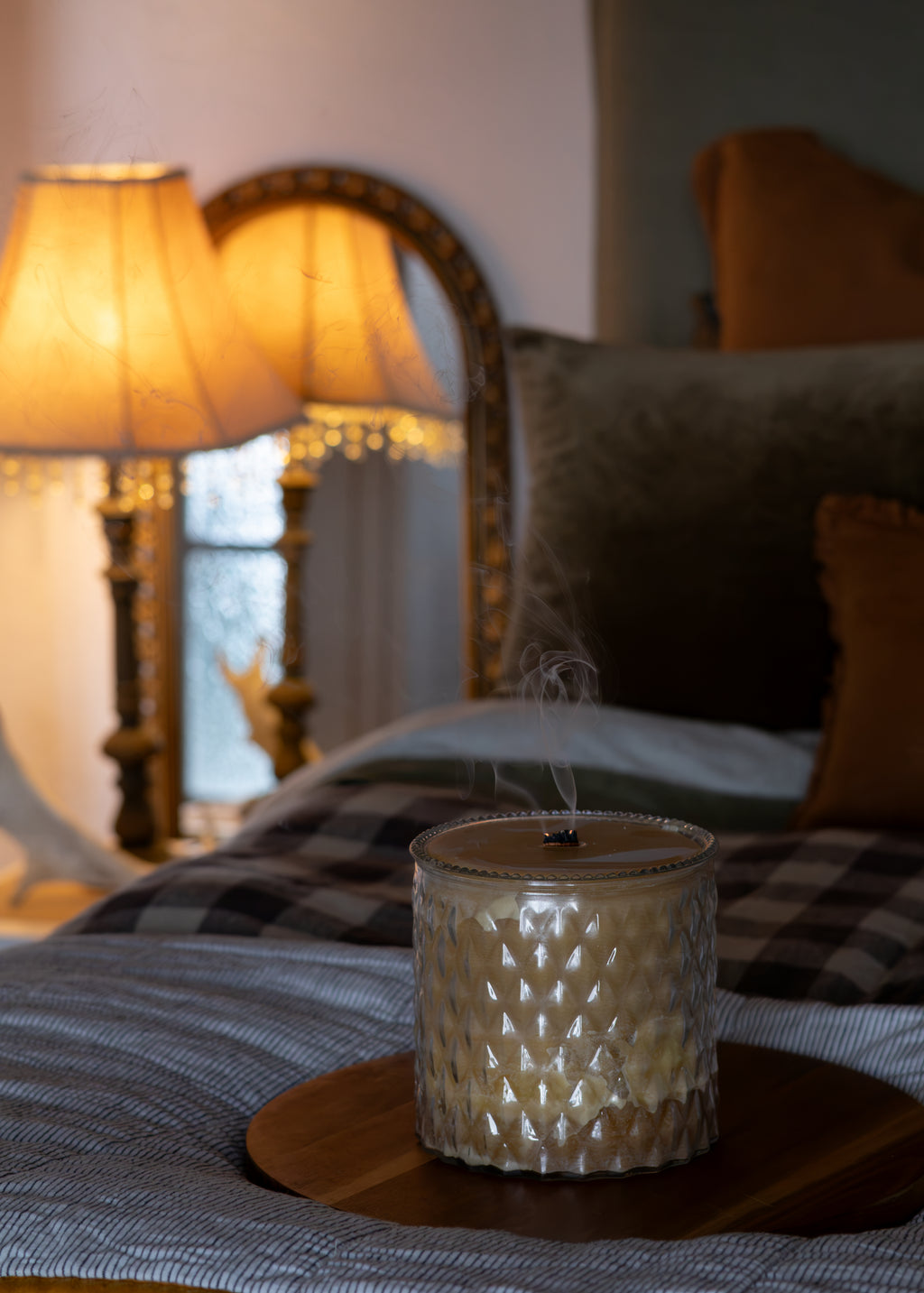 A lit scented candle in a textured glass jar sits on a round wooden tray on a bed, with a softly blurred bedroom background including a lamp, mirror, and pillows.