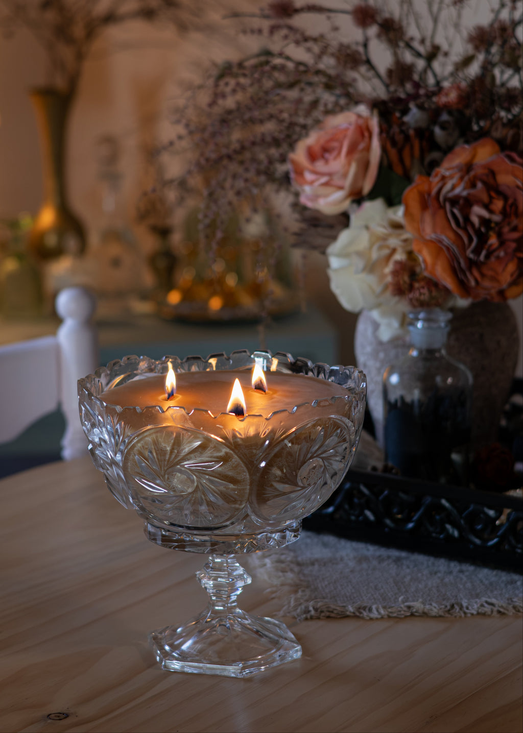 A three-wick candle in a clear, ornate glass pedestal bowl with etched floral patterns, lit on a wooden table with a warm, cozy background featuring dried flowers and a lamp.