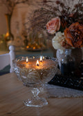 A three-wick candle in a clear, ornate glass pedestal bowl with etched floral patterns, lit on a wooden table with a warm, cozy background featuring dried flowers and a lamp.