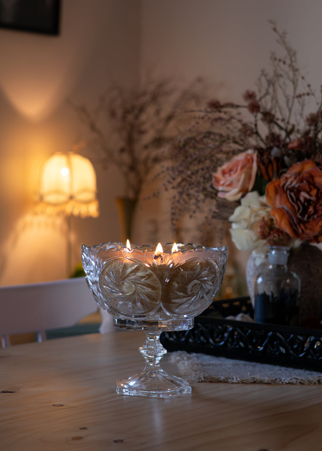 A clear cut-glass pedestal candle holder with three lit candles on a wooden table, set in a warmly lit room with a lamp and dried floral arrangements in the background.