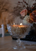A clear cut-glass pedestal candle holder with three lit candles, set on a wooden table with a blurred floral arrangement in the background.