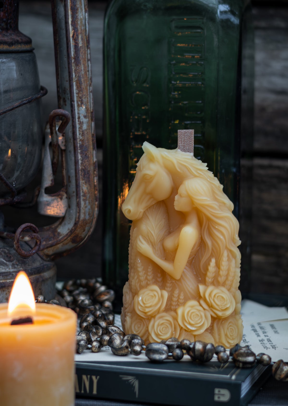 A carved beige horse figurine candle with a rose design at its hooves, displayed on a stack of vintage books next to a lit candle, a dark green glass bottle, and a rustic metal lantern against a dark wood background.