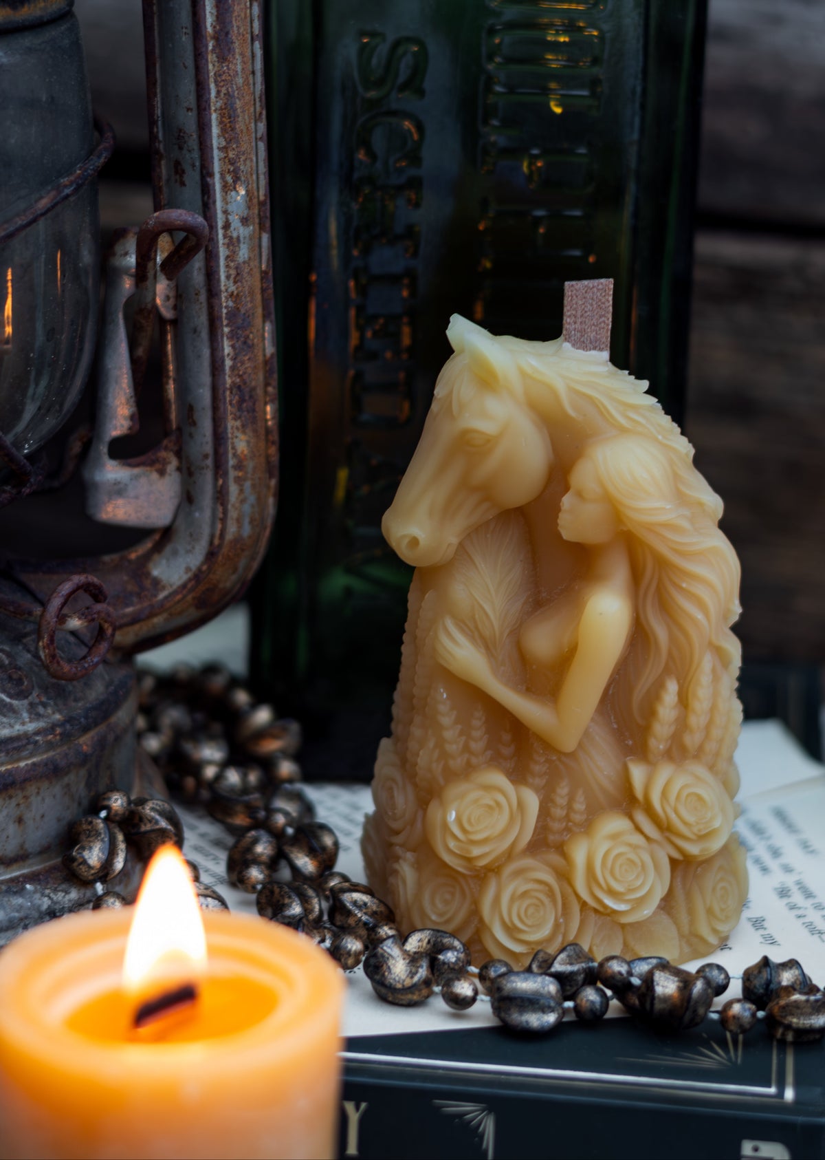 A carved cream-colored horse figurine with a woman riding it, styled on books with a lit candle, a dark green glass bottle, and a rustic lantern in the background.