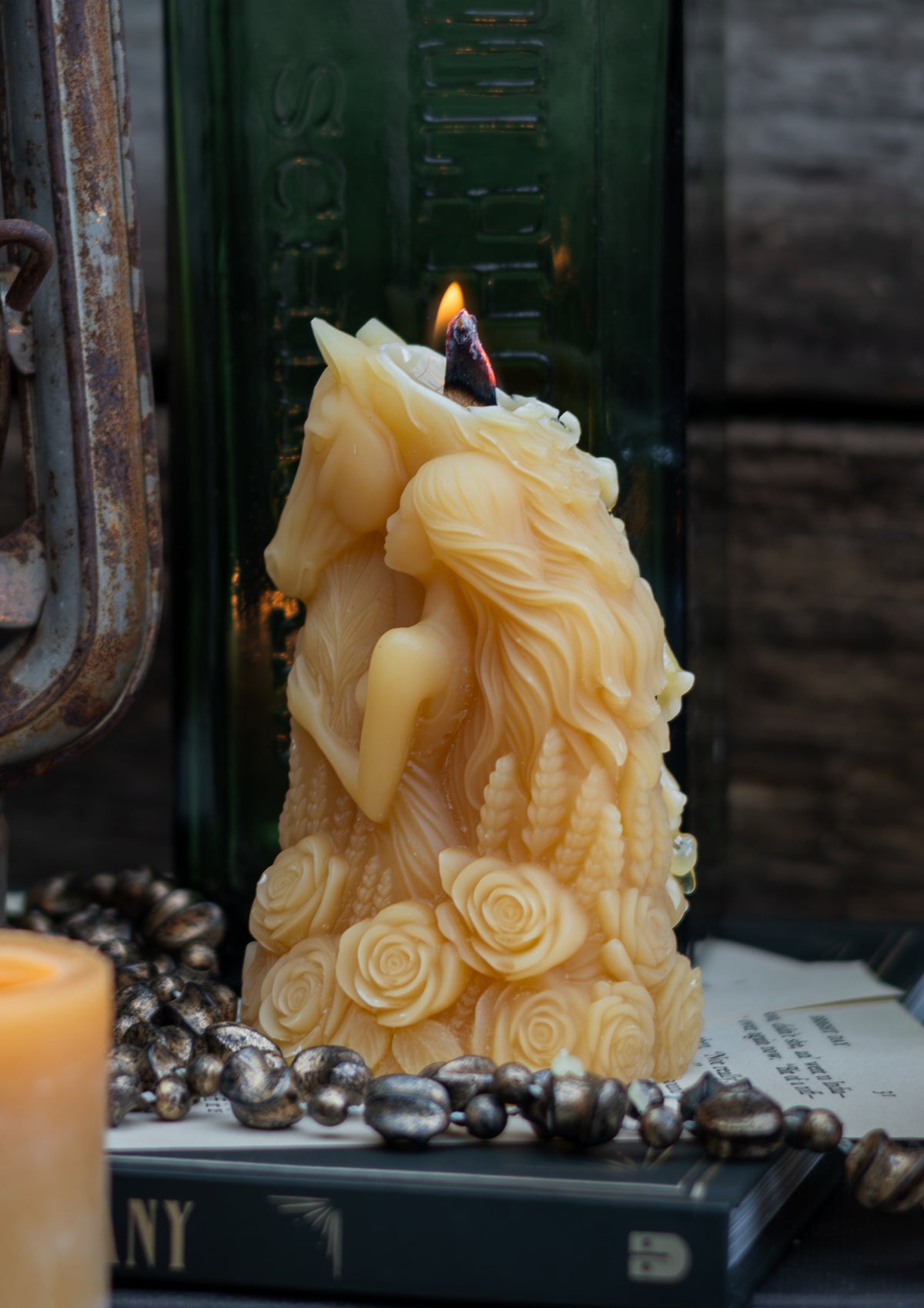 A styled rustic scene featuring a carved pure Tasmanian beeswax candle with a horse and rider design, lit with a flame, displayed on books next to a green glass bottle, an antique lantern, and another lit pillar candle against a dark wood background.